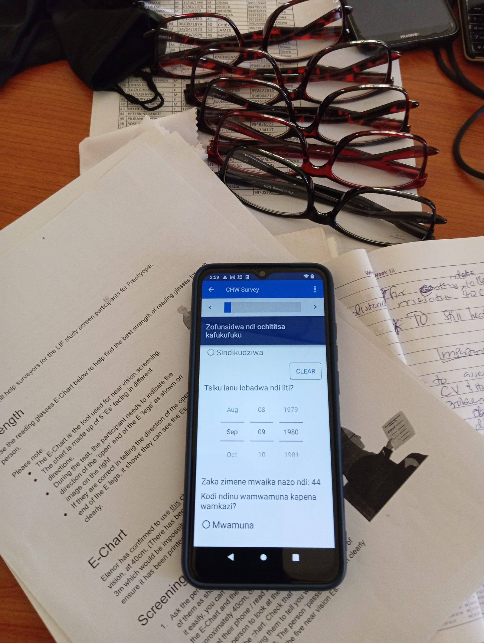 Reading glasses, a paper near-vision screening chart, and a Frontline Worker's survey phone laid out during a Reading Glasses screening session