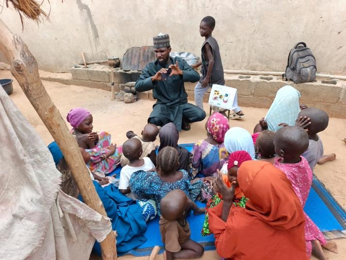 A community worker kneels on a mat demonstrating handwashing to a large group of children and carers gathered around him in a compound.