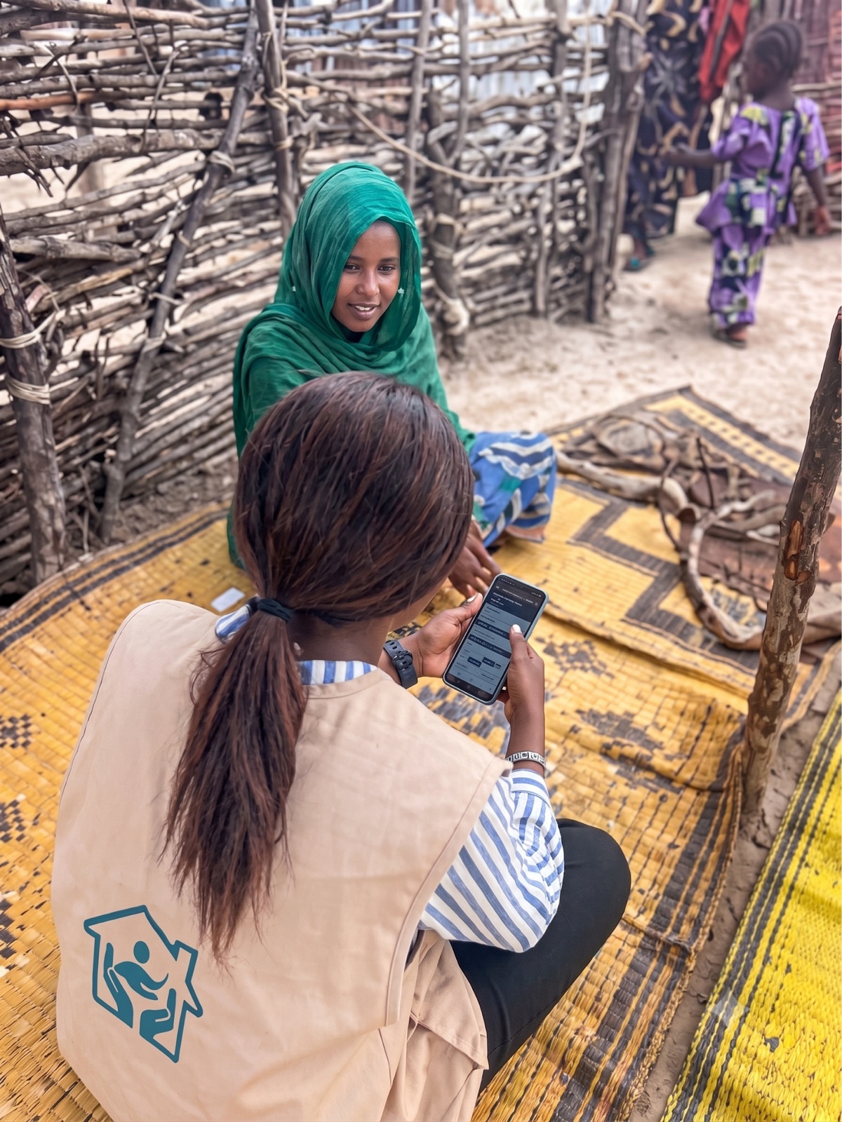 A frontline worker in a branded vest sits on a woven mat filling in a form on her phone, facing a woman in a green headscarf, inside a woven-stick compound.