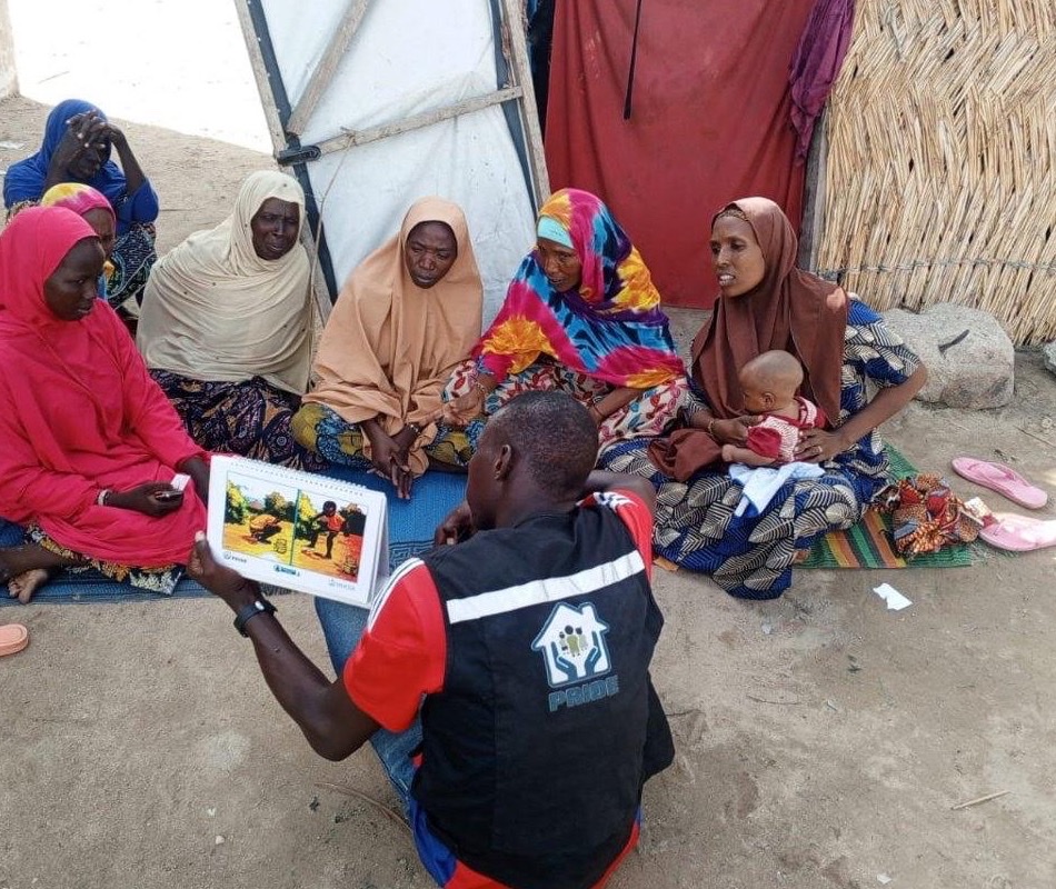 A frontline worker in a branded vest holds up an illustrated flipchart while seven women, two of them holding babies, sit on mats in a camp shelter and watch.