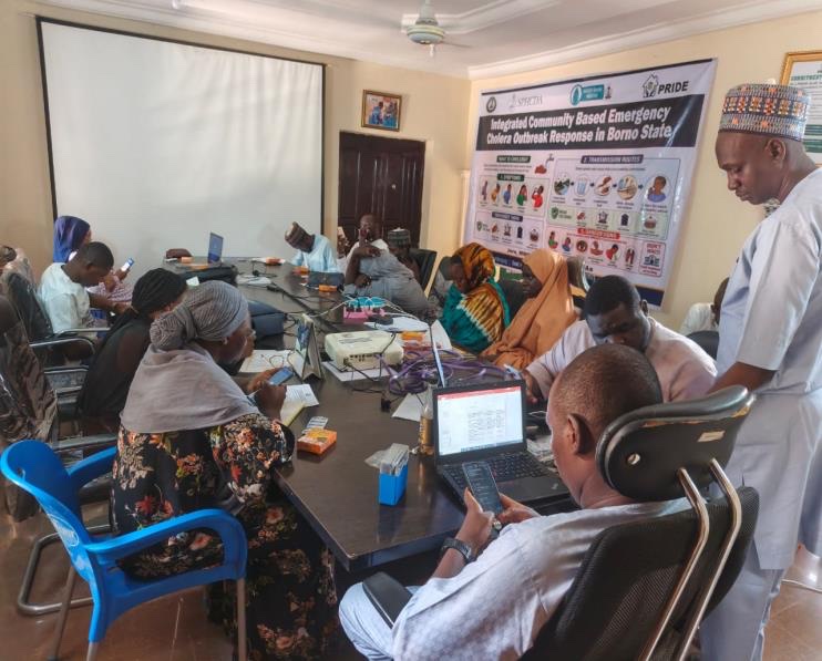 A planning room in Borno State under a banner reading Integrated Community Based Emergency Cholera Outbreak Response in Borno State. Around a long table, people work on phones and laptops, with supplies laid out in front of them.